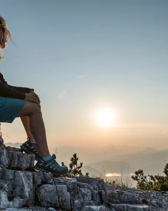 Frau sitzt auf Felsen in den Bergen und blickt ins Tal bei Sonnenuntergang